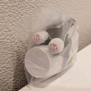 K&ouml;nnte beinhalten: Eine kleine, verpackte Katzenfigur, mit wei&szlig;en Pfoten und rosa Pfotenballen sichtbar. Die Katze ist &uuml;berwiegend grau und wei&szlig;, mit einem zylindrischen Sockel. Die Verpackung ist aus klarem Kunststoff. Der Sockel hat teilweise sichtbaren Text.