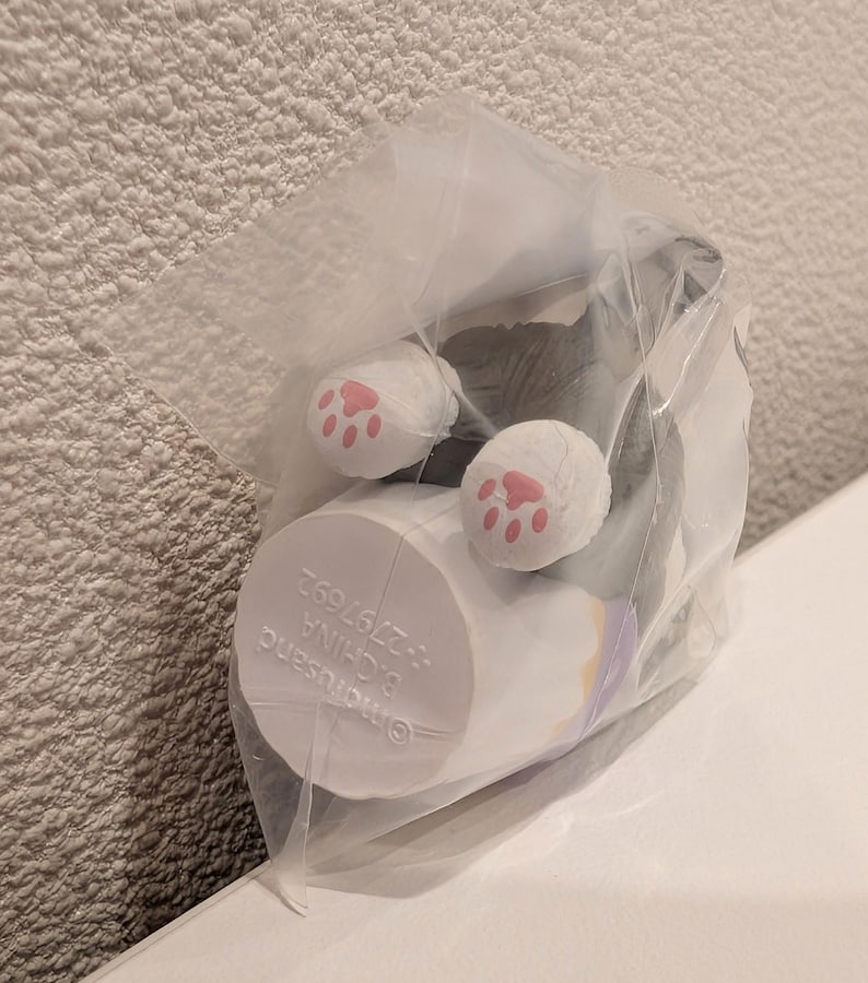 K&ouml;nnte beinhalten: Eine kleine, verpackte Katzenfigur, mit wei&szlig;en Pfoten und rosa Pfotenballen sichtbar. Die Katze ist &uuml;berwiegend grau und wei&szlig;, mit einem zylindrischen Sockel. Die Verpackung ist aus klarem Kunststoff. Der Sockel hat teilweise sichtbaren Text.