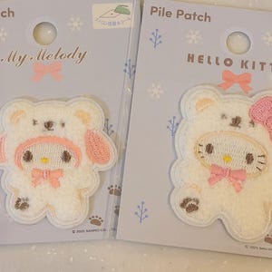 May include: Two white pile patches on individual display cards. The left patch features My Melody wearing a white bear hood with pink accents. The right patch features Hello Kitty wearing a white bear hood with a pink bow. Both have snowflake designs.