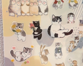May include: A sheet of stickers featuring various cartoon cat illustrations. The stickers depict cats in different poses, some holding flowers or wearing outfits. The color palette includes shades of gray, brown, white, and black.