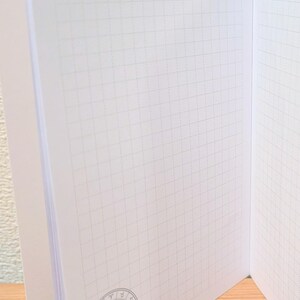 May include: Open notebook with grid paper pages. The notebook has a decorative border at the top with the word "mofusand". A circular illustration of two cat-like characters is in the lower corner. The pages are white.