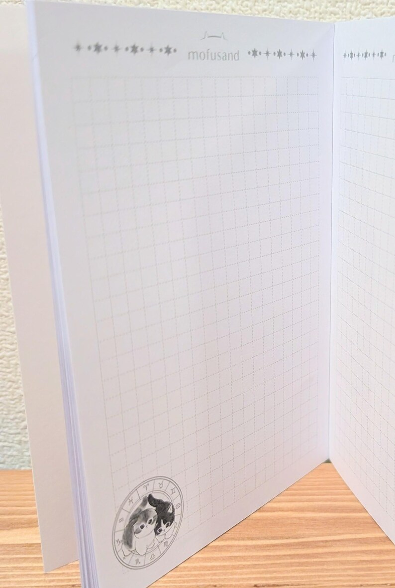 May include: Open notebook with grid paper pages. The notebook has a decorative border at the top with the word "mofusand". A circular illustration of two cat-like characters is in the lower corner. The pages are white.