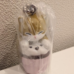 K&ouml;nnte beinhalten: Katzenfigur im Cupcake-Design, in klarem Kunststoff verpackt. Die Katze hat ein Gold- und Wei&szlig;-Farbschema, mit einer rosa Cupcake-Basis und einem braunen Zuckerguss-Detail. Ein silberfarbener Anh&auml;nger ist oben an der Figur befestigt.