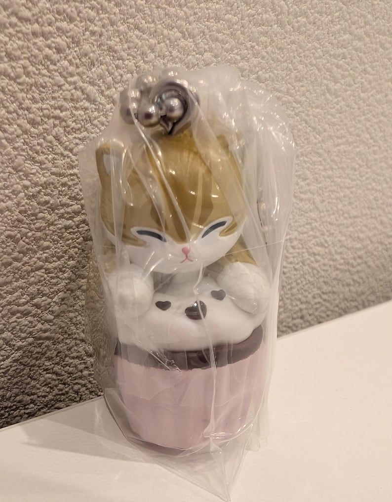 K&ouml;nnte beinhalten: Katzenfigur im Cupcake-Design, in klarem Kunststoff verpackt. Die Katze hat ein Gold- und Wei&szlig;-Farbschema, mit einer rosa Cupcake-Basis und einem braunen Zuckerguss-Detail. Ein silberfarbener Anh&auml;nger ist oben an der Figur befestigt.