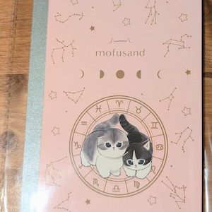 May include: A pink notebook featuring two cartoon cats within a zodiac wheel design. The notebook has a silver spine and is decorated with constellations and the text "mofusand". The brand name "@mofusand" is also visible.