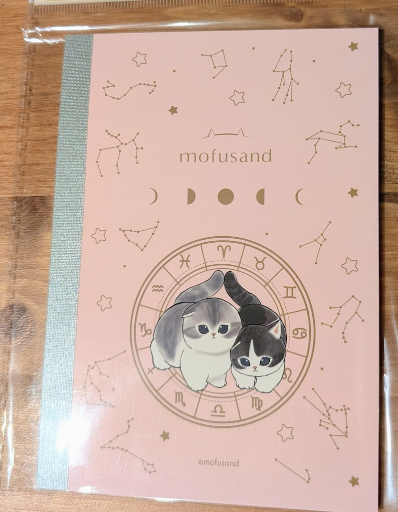 May include: A pink notebook featuring two cartoon cats within a zodiac wheel design. The notebook has a silver spine and is decorated with constellations and the text "mofusand". The brand name "@mofusand" is also visible.