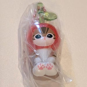 May include: A small figurine of a brown tabby cat wearing a red cherry-shaped hood with a green stem and leaf on top. The cat has large black eyes, white paws with pink paw pad markings, and is encased in a clear, crinkled plastic protective bag.
