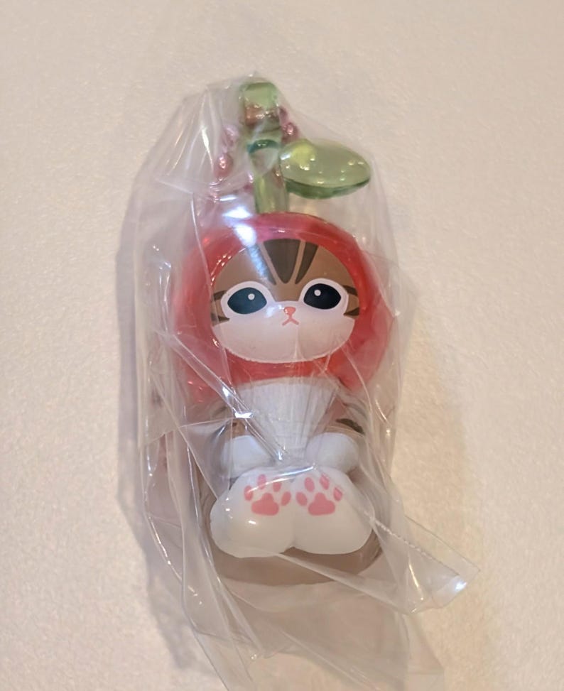 May include: A small figurine of a brown tabby cat wearing a red cherry-shaped hood with a green stem and leaf on top. The cat has large black eyes, white paws with pink paw pad markings, and is encased in a clear, crinkled plastic protective bag.