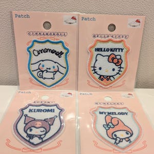 May include: Four embroidered shield-shaped patches featuring Sanrio characters Cinnamoroll, Hello Kitty, Kuromi, and My Melody. Each patch is displayed on a light pink backing card with the text Patch and an iron-on instruction icon included.