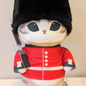 May include: A plush cat toy dressed as a British royal guard, wearing a tall black fuzzy bearskin hat and a bright red tunic with white buttons and a belt. The cat has grey tabby markings on its face and body, large dark eyes, and holds a grey rifle.