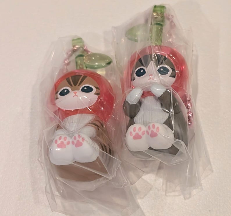 May include: Two cat figurines in clear plastic packaging, each wearing a red hood with a green leaf accent. One cat is brown tabby, the other is black and white. Both have white paws with pink paw pads and are attached to a pink ball chain keychain.