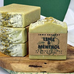 May include: Four bars of handmade soap, stacked on a wooden board. The soap is light green with a white top and speckled with seeds. One bar has a brown paper label that reads "Lime & Menthol" and "Handmade Soap in Des Moines, IA."