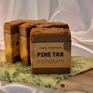 May include: Handmade Pine Tar soap bars from Iowa Soapery. The brown soap is wrapped in paper with the text "Pine Tar" and "Handmade Soap in Des Moines, IA". The soap is displayed on a floral patterned surface.