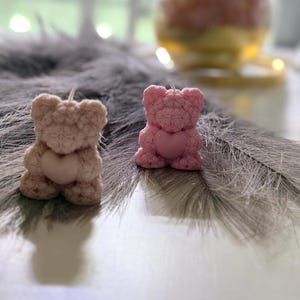 May include: Two small pink teddy bear candles with a heart design on their chests. The candles are sitting on a grey fluffy surface.