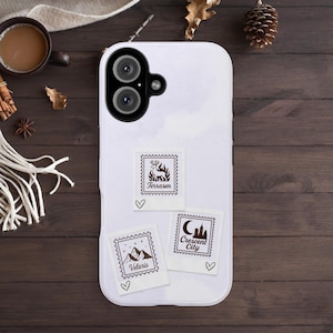 May include: White phone case with three decorative postage stamp designs. The stamps feature illustrations of a deer, mountains, and a city skyline, each accompanied by a heart symbol and text: 'Terrasen', 'Velaris', and 'Crescent City'.