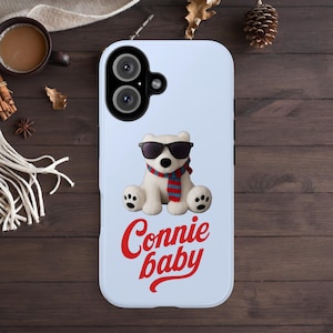Puede incluir: Funda de teléfono azul claro con un oso polar blanco que lleva gafas de sol y una bufanda roja y azul. El texto "Connie baby" está escrito en cursiva roja debajo. La funda tiene un recorte para cámara de doble lente.