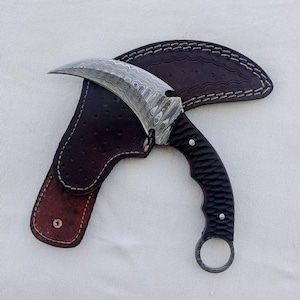 May include: A curved karambit knife with a patterned blade and a textured black handle. The knife rests in a brown leather sheath with white stitching. The blade has a unique wave pattern.