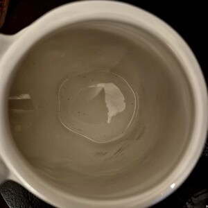 May include: A white ceramic mug with a handle, viewed from above. The interior of the mug is a light beige color, with a few dark specks visible. The mug is empty.