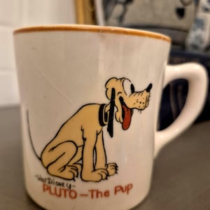 Puede incluir: Taza de cerámica blanca con Pluto, el perro de dibujos animados de Disney. La taza tiene un borde y un asa marrones. Pluto está representado sentado con la lengua fuera. El texto "Walt Disney - Pluto - The Pup" está impreso en la taza.