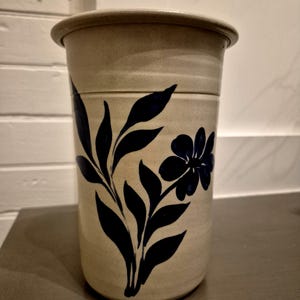 May include: A beige ceramic jar with a lid, featuring a dark blue floral design. The jar has a cylindrical shape and a slightly textured surface. The floral pattern includes leaves and a flower.