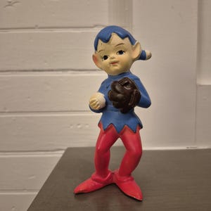 May include: A vintage ceramic figurine of a baseball-playing elf. The elf is dressed in a blue top with a red jagged hem, red tights, and a brown baseball glove. The elf has a blue cap and a neutral skin tone.