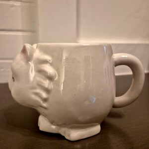 May include: A white ceramic mug shaped like a cat. The mug features a detailed cat head on one side, with a handle on the other. The cat's ears have pink accents. The mug sits on a rectangular base.