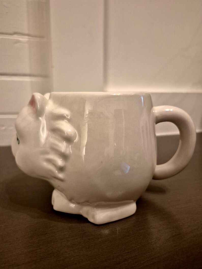 May include: A white ceramic mug shaped like a cat. The mug features a detailed cat head on one side, with a handle on the other. The cat's ears have pink accents. The mug sits on a rectangular base.