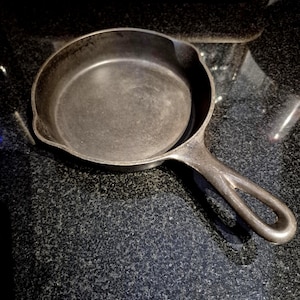 May include: A seasoned cast iron frying pan with a single handle. The pan is dark grey with a slightly textured surface, showing signs of use. The handle has a loop for hanging. A classic kitchenware item.