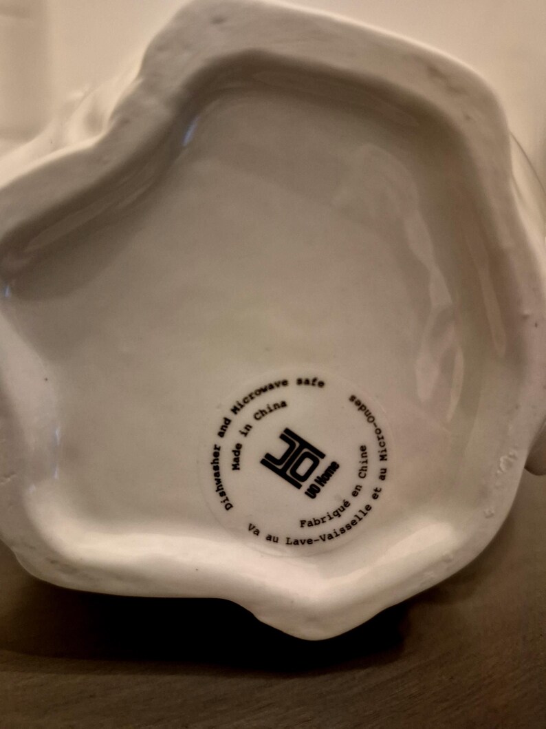 May include: A white ceramic serving dish with a rounded shape. The bottom of the dish has a circular sticker with text that reads "Dishwasher and Microwave safe Made in China UO Home Va au Lave-vaisselle et au Micro-Onde Fabriqu&eacute; en Chine."