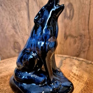 May include: A ceramic figurine of a howling wolf, glazed in a glossy, dark blue with streaks of lighter blue. The wolf is depicted in a seated position, head raised and mouth open, on a rounded base.