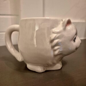 May include: A white ceramic mug shaped like a cat's head. The mug has a handle and the cat's face is detailed with eyes, ears, and a nose. The mug is sitting on a dark surface.