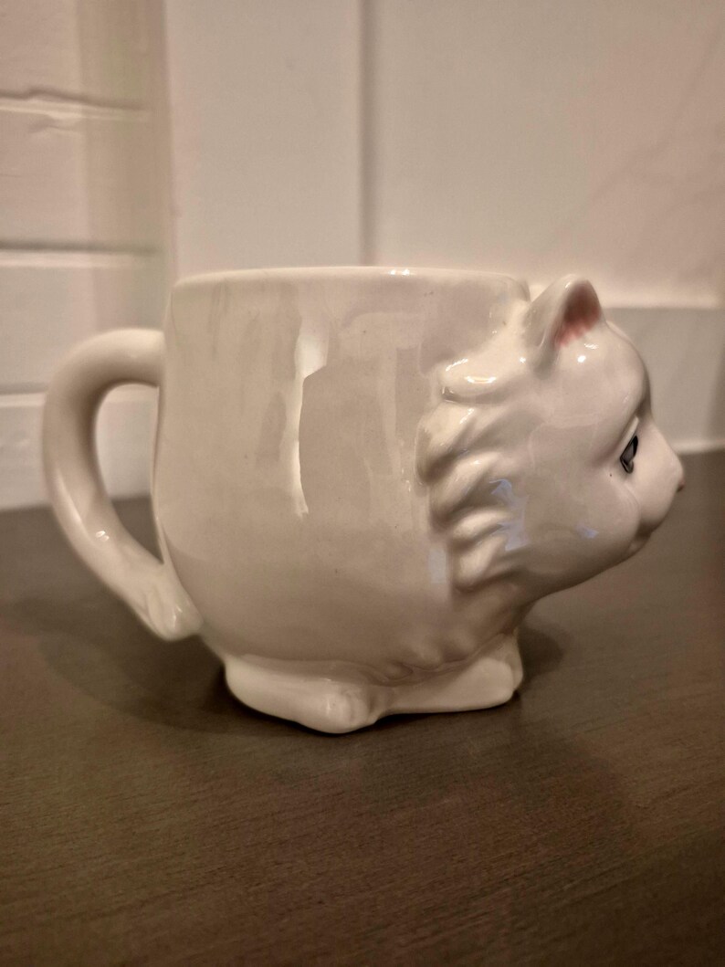 May include: A white ceramic mug shaped like a cat's head. The mug has a handle and the cat's face is detailed with eyes, ears, and a nose. The mug is sitting on a dark surface.