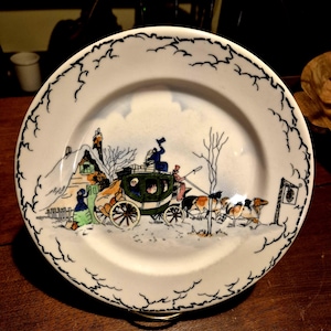 Puede incluir: Un plato decorativo blanco con un borde festoneado negro. El plato presenta una escena invernal con un carruaje tirado por caballos, personas y un edificio. La escena está en tonos de verde, azul, marrón y blanco.