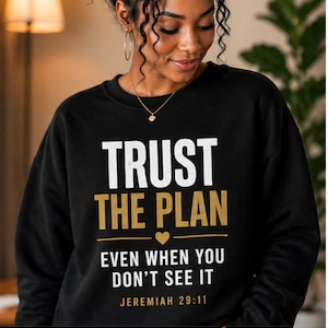 Pode incluir: Camisola preta com a frase "TRUST THE PLAN" em letras brancas e douradas, juntamente com o versículo bíblico Jeremias 29:11. O design inclui um símbolo de coração. Ideal para vestuário e acessórios.