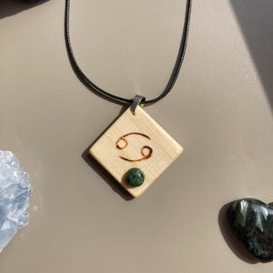 May include: A wooden pendant necklace with the Cancer zodiac symbol engraved on a diamond-shaped wooden pendant. A small, round, green stone is set below the symbol. The pendant hangs from a black cord.