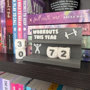 Puede incluir: Un rastreador de fitness gris con las palabras "WORKOUTS THIS YEAR" y un icono de mancuerna. Muestra los números 30 y 72 en dados blancos, indicando el número de entrenamientos. El rastreador está en una estantería con libros.
