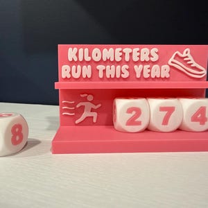 May include: A pink running tracker with the text "KILOMETERS RUN THIS YEAR" and a running shoe graphic. It includes three dice displaying the numbers 274, and a single die showing 18. The tracker is designed to track running distance.