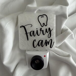 May include: A white night light with a square, textured top featuring a tooth graphic and the words "Fairy cam" in black script. The night light has a black lens and a red dot.