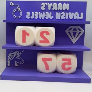 Op de afbeelding: Een paarse displayplank met twee niveaus, met witte dobbelstenen met roze cijfers, een diamant, een eenhoorn en de tekst "MARY'S JEWEL TEMPLE" in het wit. De bovenste plank heeft een bom- en sterontwerp.