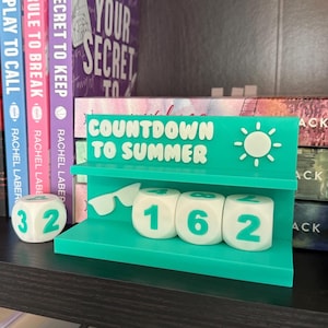 May include: A teal countdown calendar with the words "COUNTDOWN TO SUMMER" and a sun graphic. The calendar has three dice displaying the numbers 1, 6, and 2. A separate die shows the number 32. Books are in the background.