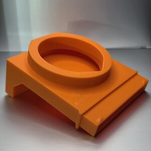 May include: Vibrant orange plastic component with a circular top and a rectangular base. The object's design is distinctive, possibly a tool or accessory. The surface is smooth, and the colour is uniform throughout.