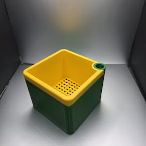 May include: A square, two-tone container with a green exterior and a yellow interior. The rim and a small circular opening are also yellow. The interior has a grid-like pattern. The container is likely 3D-printed.