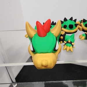 Può includere: Una figura di Bowser Jr. stampata in 3D, con testa verde, corna e muso gialli e cresta rossa. La figura fa parte di un set, con altri pezzi di guscio verdi e gialli a spillo visibili.