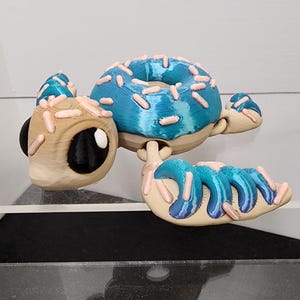 May include: A quirky 3D-printed turtle ornament with a donut shell. The turtle has a blue donut with pink sprinkles, a tan head and limbs, and black eyes. This unusual design would make a fun gift or decorative item for a home.
