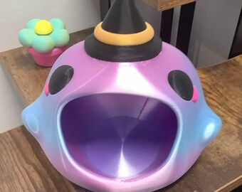 May include: A quirky, iridescent purple, blue, and pink creature-shaped container with a witch's hat. The container has a large opening and black eyes. A small flower-shaped object is in the background. The container is on a wooden surface.