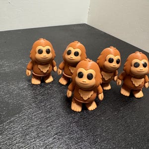 Puede incluir: Cinco pequeñas figuras de monos marrones con grandes ojos negros y detalles de pelaje marrón claro. Cada figura tiene un pecho y pies de color marrón claro. Los monos están sobre una superficie negra.