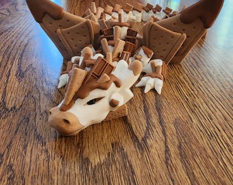 May include: A whimsical, brown and white dragon sculpture made of what appears to be graham crackers, marshmallows, and chocolate. The dragon has large wings and a detailed head, with a textured wooden table in the background.