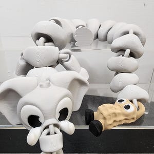 Puede incluir: Un juguete de elefante articulado gris claro con orejas grandes y un cuerpo segmentado. El elefante tiene ojos grandes y una trompa. También hay una figura en forma de cacahuete beige con pies y ojos negros.