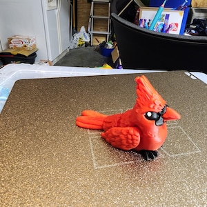 May include: A vibrant red cardinal figurine with black accents on its beak and feet. The bird has a detailed wing and tail design, and is set against a textured, gold-colored surface. The figurine is a decorative item.