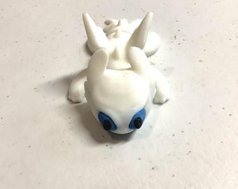 May include: A small white figurine of a dragon-like creature with large pointed ears, a smooth body, and textured ridges along its back. The creature features prominent blue eyes with black pupils, and it is positioned in a prone, resting posture.